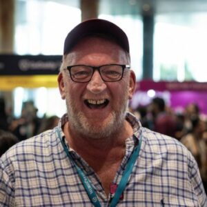 A smiling older man with glasses wearing a maroon cap and a checkered shirt with a lanyard, standing in a crowded indoor area.