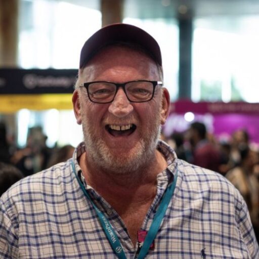 A smiling man wearing glasses, a cap, and a checked shirt at a busy indoor event.