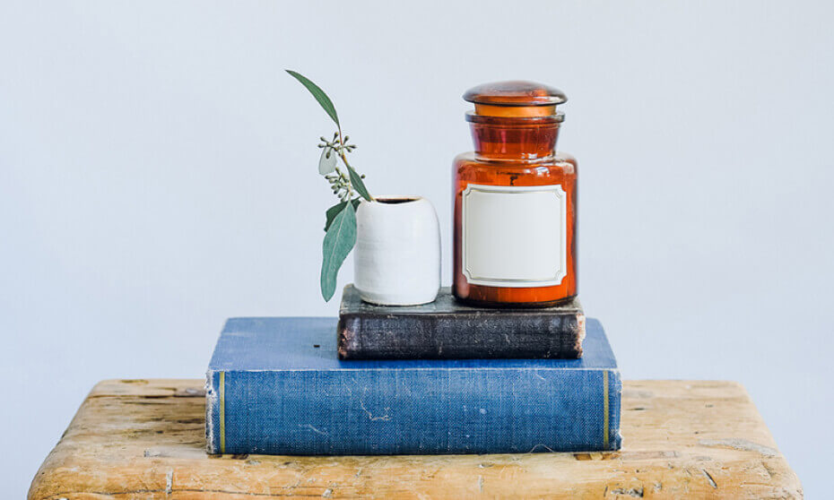 An old brown apothecary jar on stacked vintage books beside a small ceramic vase with a plant sprig, on a wooden surface against a plain background